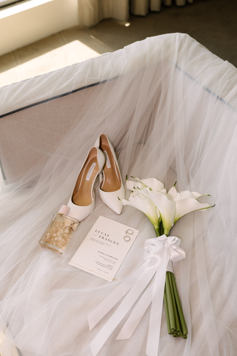 A styled bridal scene with a pair of white pointed-toe heels, a bouquet of white calla lilies tied with a white ribbon, a small card, and a decorative bottle arranged on white tulle fabric.