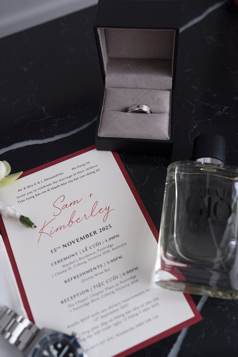 A wedding invitation card with red borders and script text lies on a dark marble surface next to an open ring box containing a silver band, a perfume bottle, and a watch.