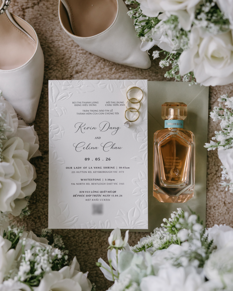 Wedding invitation card with rings and perfume bottle surrounded by white flowers on a textured surface