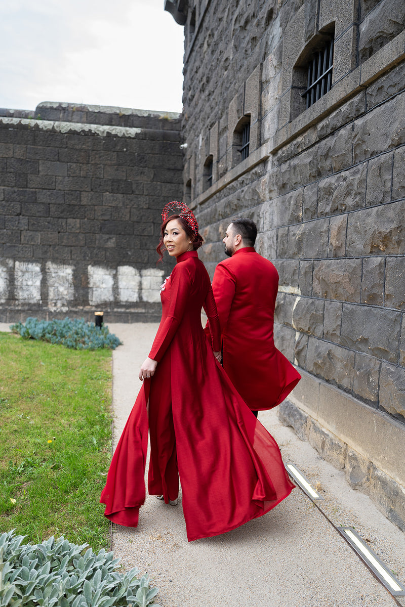 A couple in matching bright red Vietnamese Ao Dai walking along a stone path beside a castle wall. The woman wears a long red traditional outfit with a headpiece, and the man wears a red tailcoat.